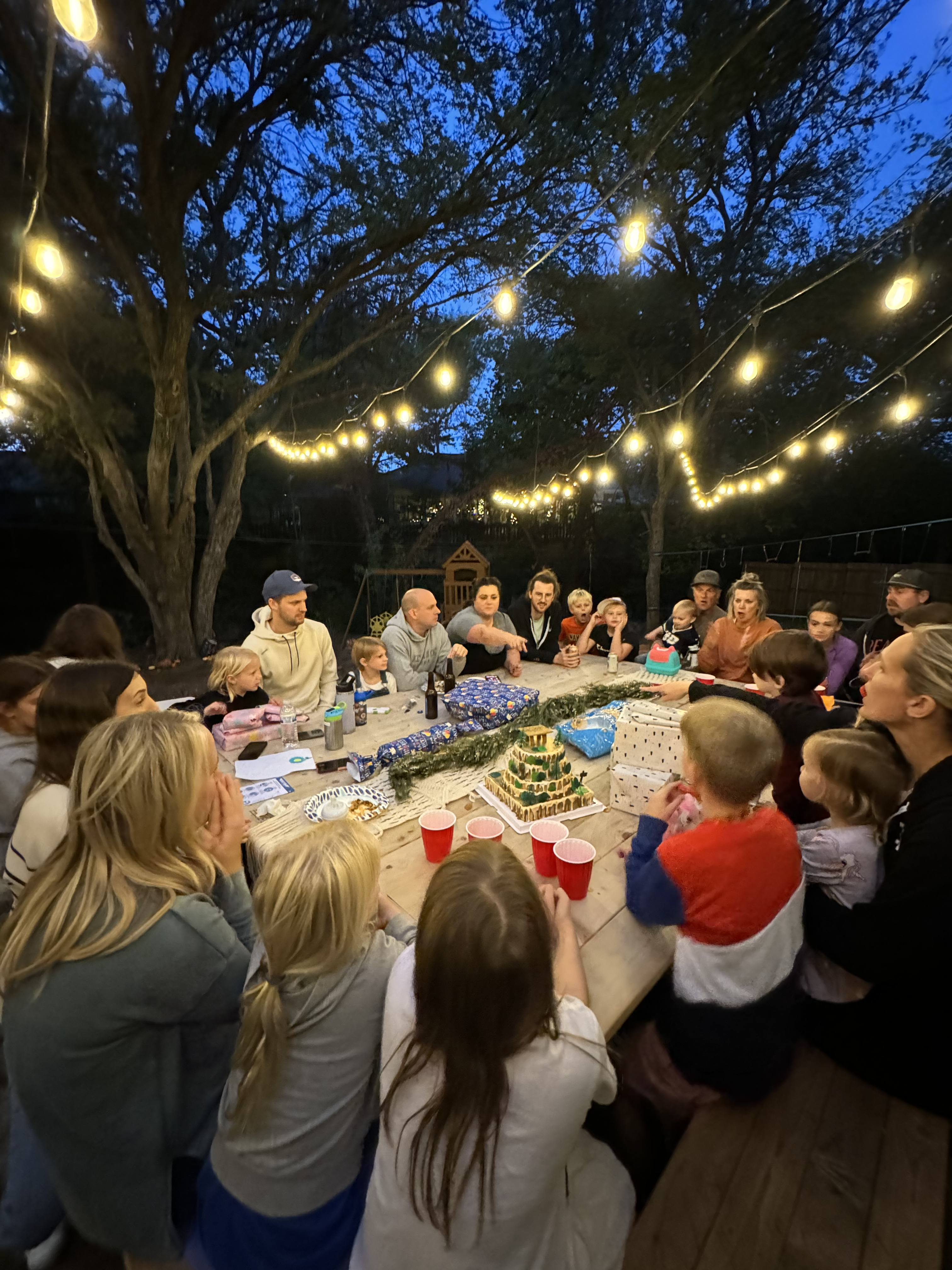 Family gathered around a large wooden table on the patio at dusk with lights strung up above.